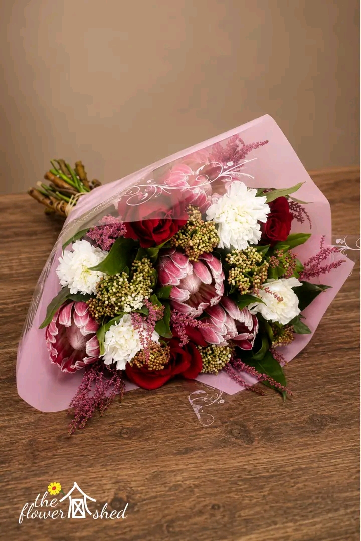 A bouquet of red roses, white carnations, and pink protea flowers wrapped in pink cellophane